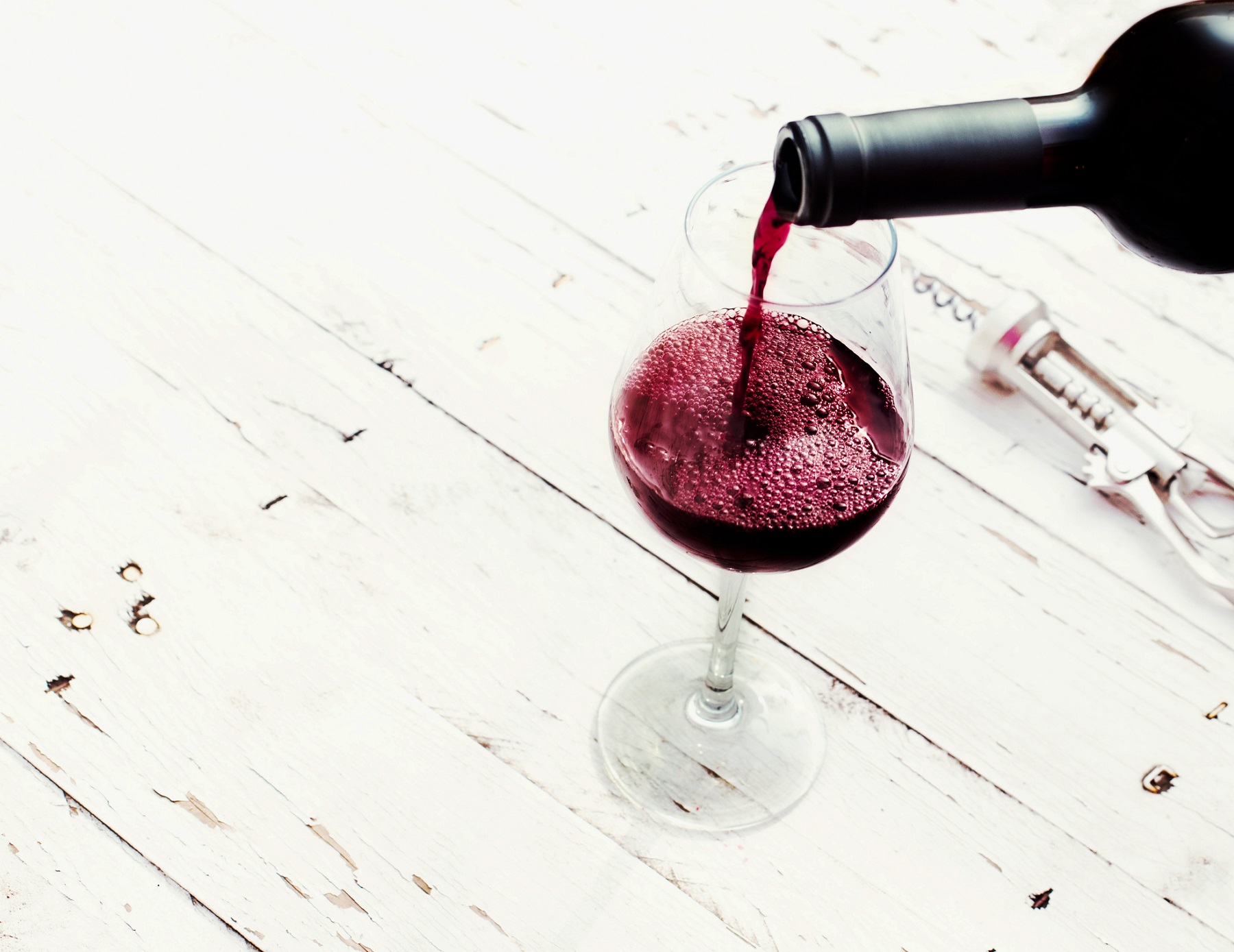Pouring red wine from bottle in wine glass on vintage white wooden table with copyspace.